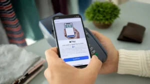 Pessoa realizando pagamento usando Apple Pay no iPhone, com um leitor de cartão no ambiente de casa, ao lado de uma carteira e uma planta.
