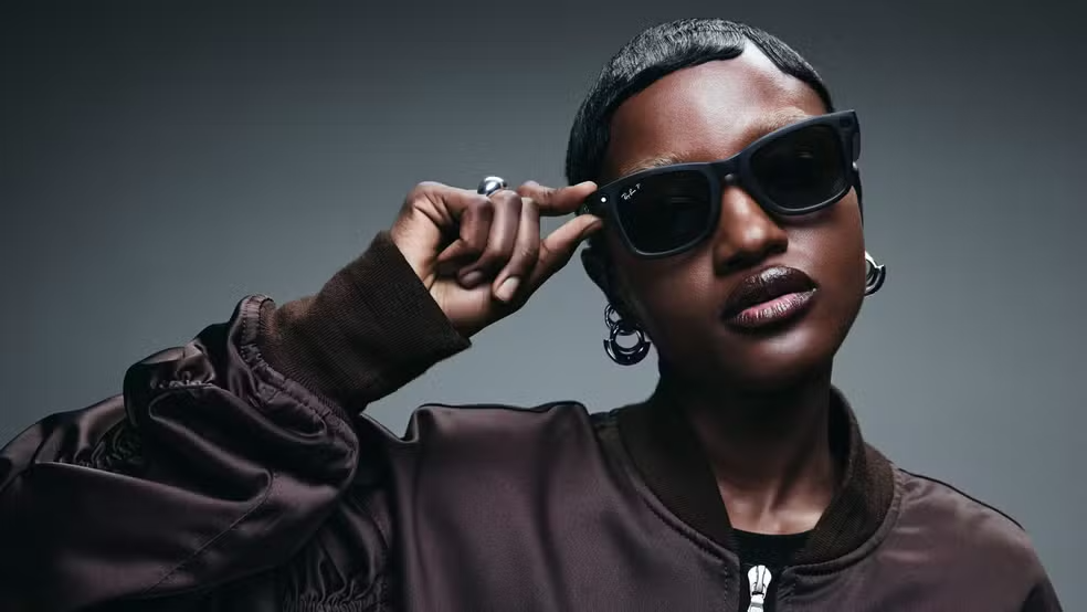 Mulher estilosa com pele escura usando óculos de sol escuros, jaqueta marrom e cabelo curto, posando com atitude contra fundo cinza.