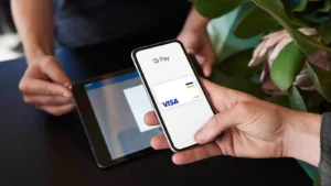 Pessoa realizando pagamento via Google Pay no celular, com cartão Visa exibido na tela, em ambiente com plantas ao fundo.