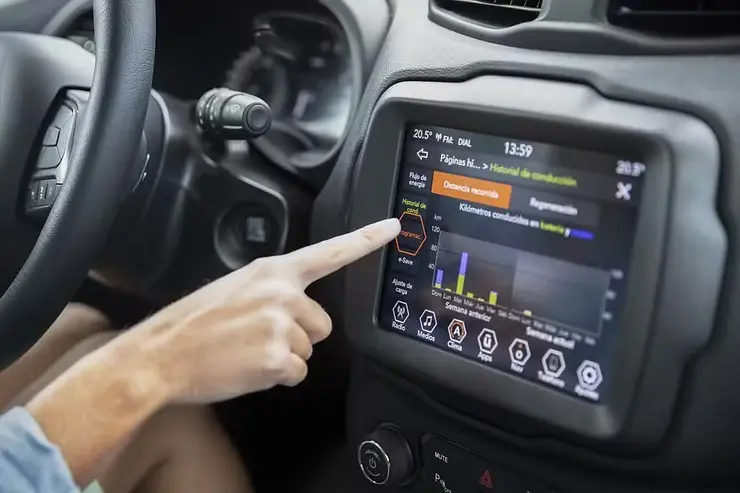 Pessoa usando o painel multimídia do carro, com tela exibindo informações de condução e gráficos, enquanto mantém a mão no volante e aponta para o visor. Close no interior do veículo.