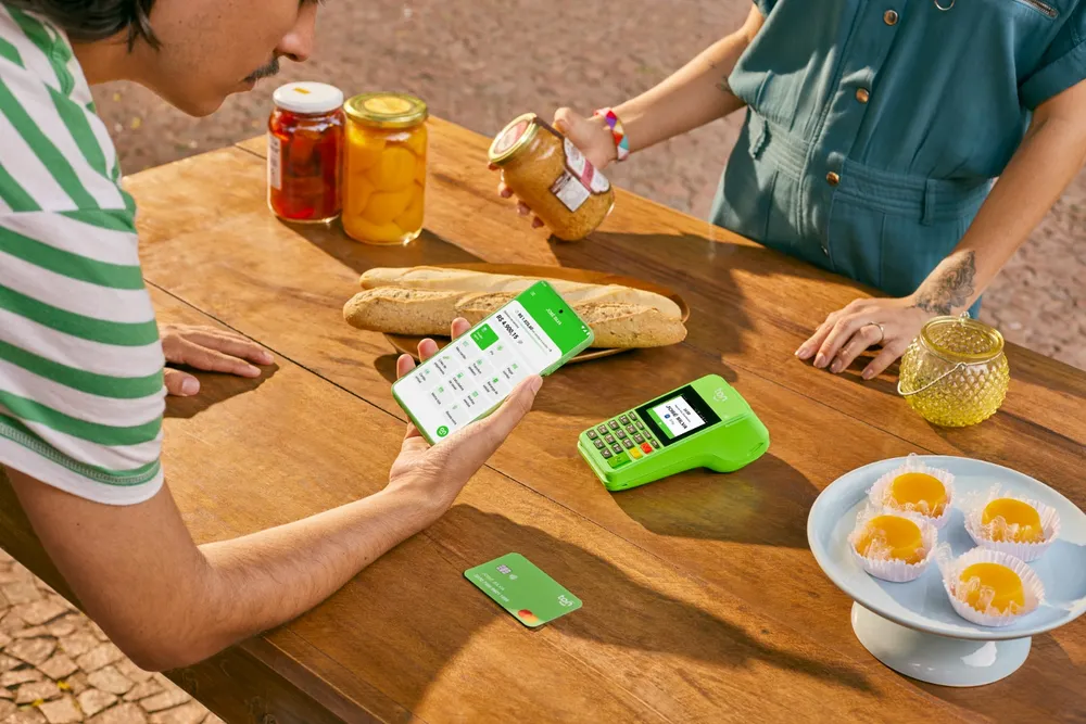 Pessoas usando maquininha de cartão e celular para pagamento em mesa de madeira com alimentos e potes, enquanto preparam um lanche ao ar livre.